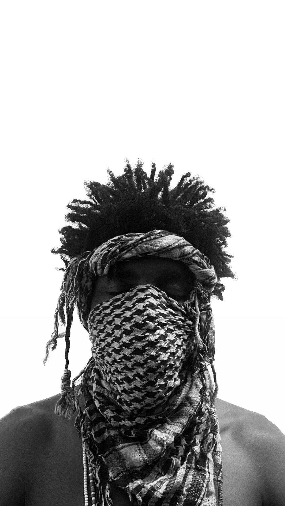 Person with head covering and patterned face mask, hair styled in an afro, wearing sunglasses, against a white background.