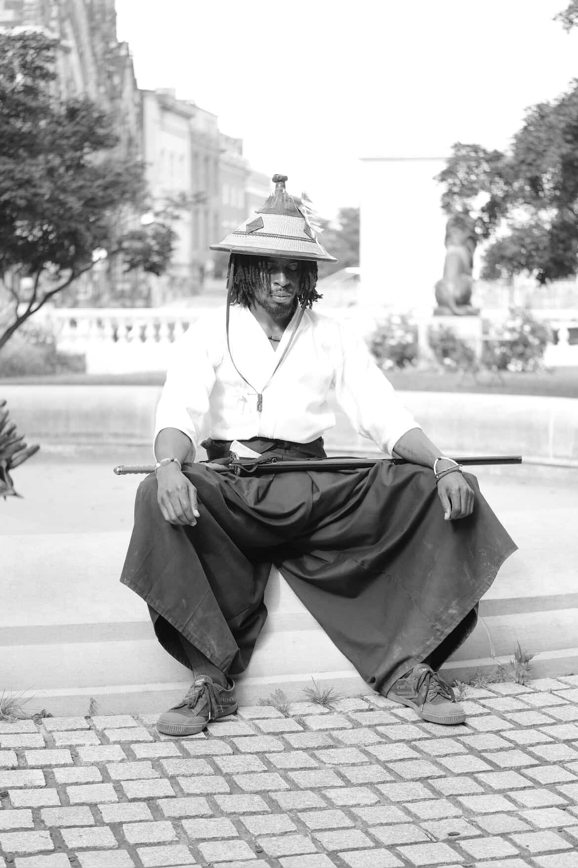 Person sitting on a bench wearing a wide-brimmed hat and white shirt, with buildings and trees in background.