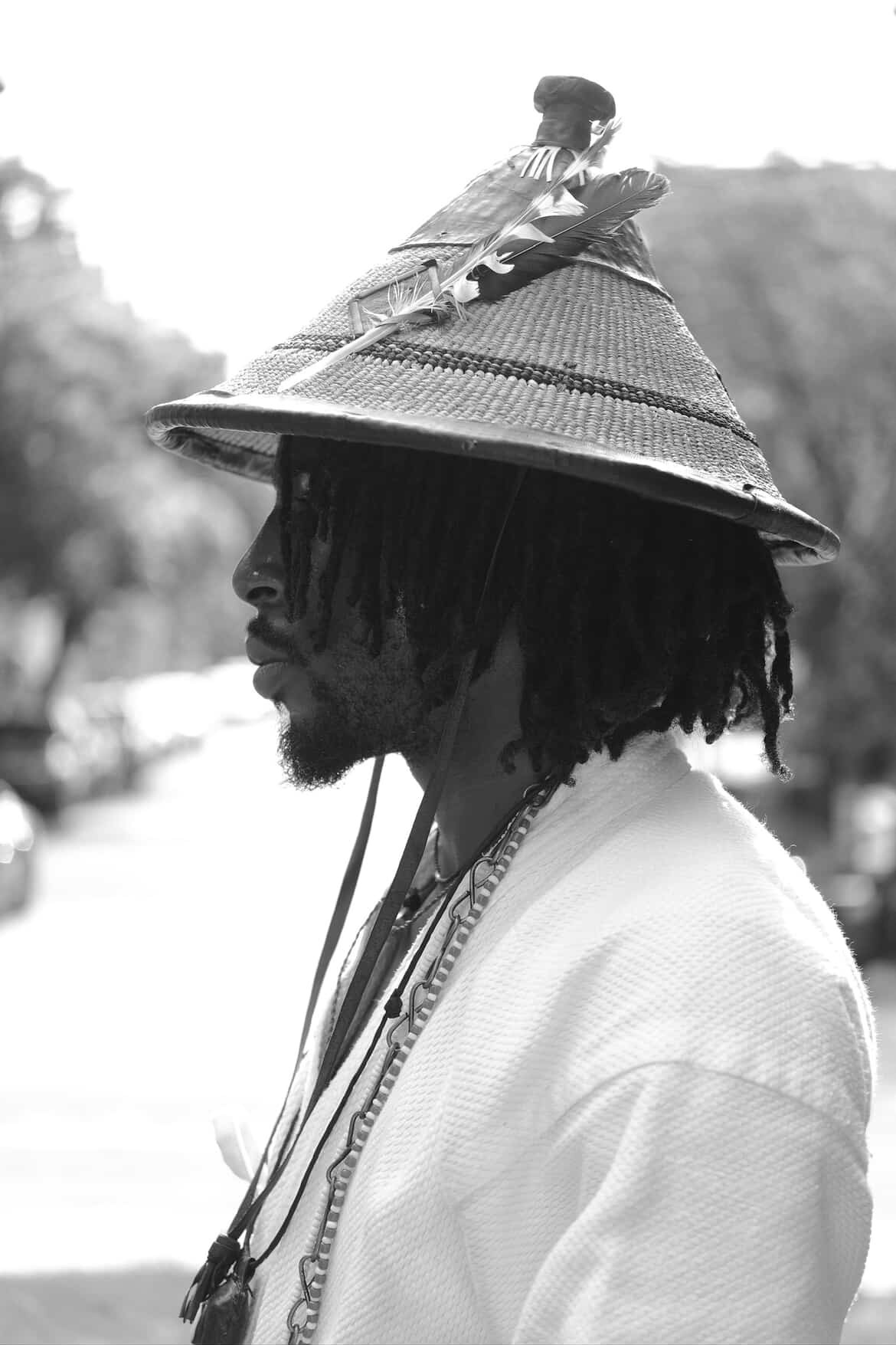 Profile of a person wearing a large conical hat with a small figure on top, outdoors, in black and white.