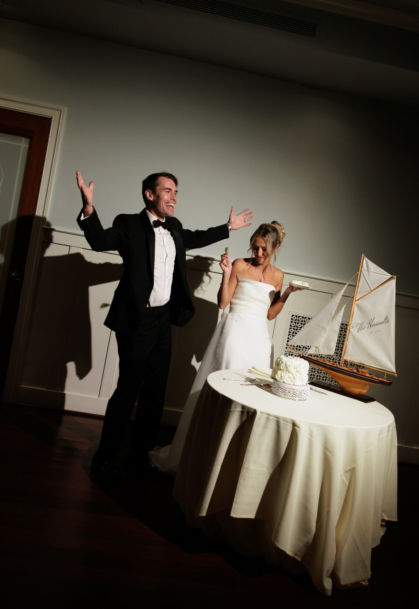 Man in tuxedo and woman in white dress stand near table with model ship, woman reads paper, man gestures outward.