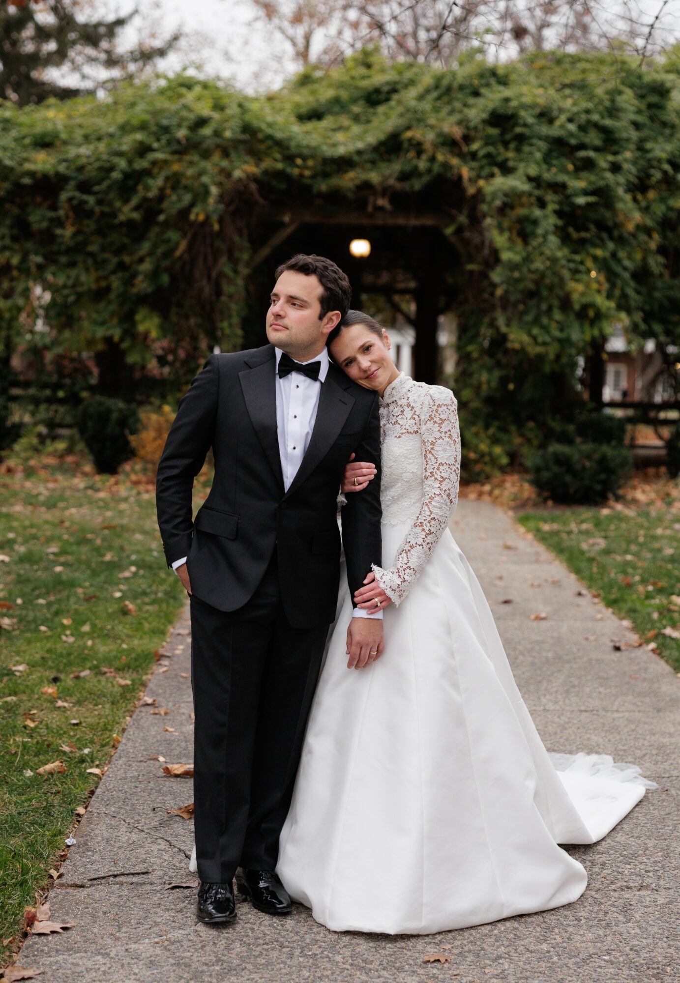 Bride and groom standing on a path outdoors, with greenery and an archway in the background.