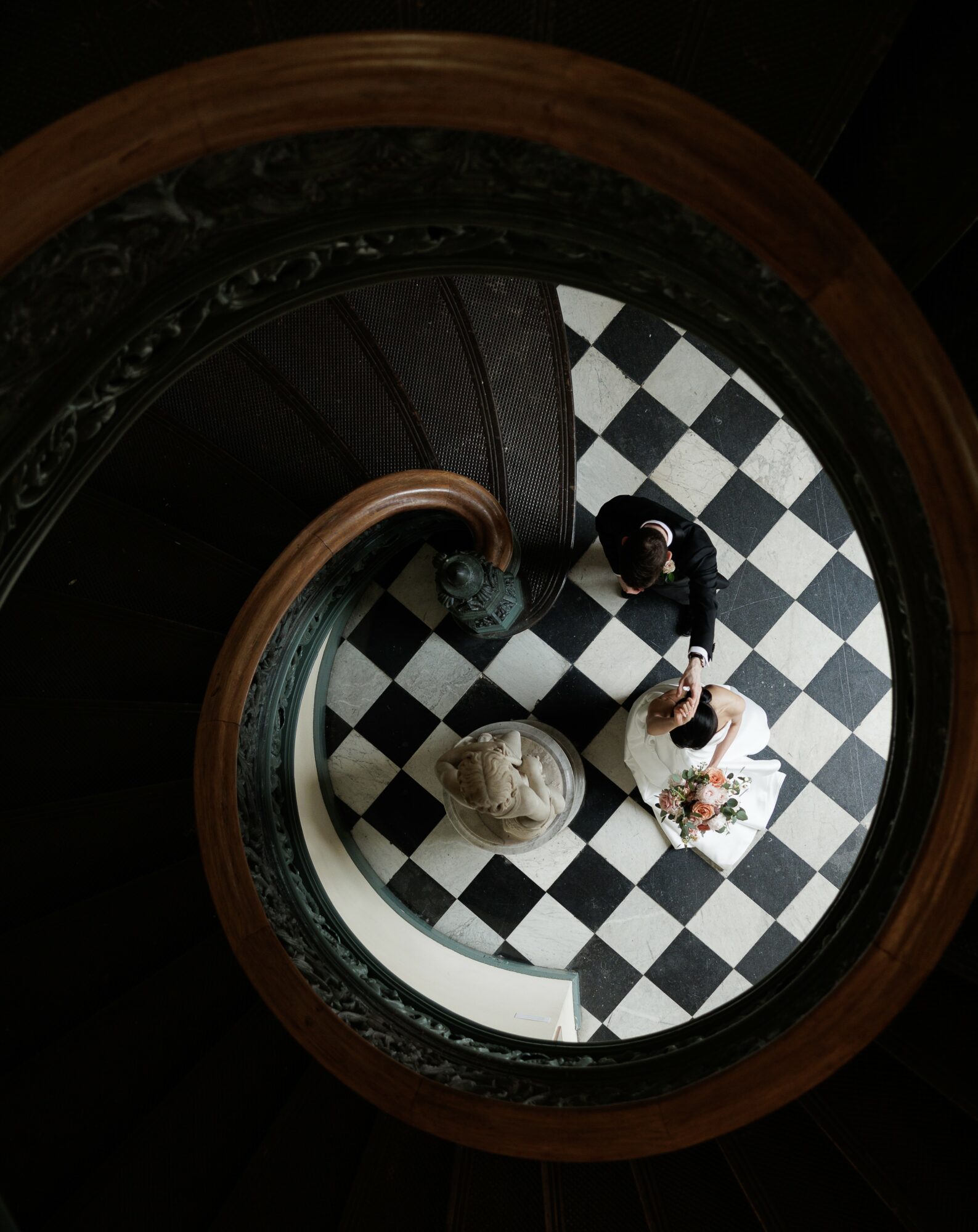 Two people stand on a black and white checkered floor, viewed from above, near a curved staircase with a decorative railing.