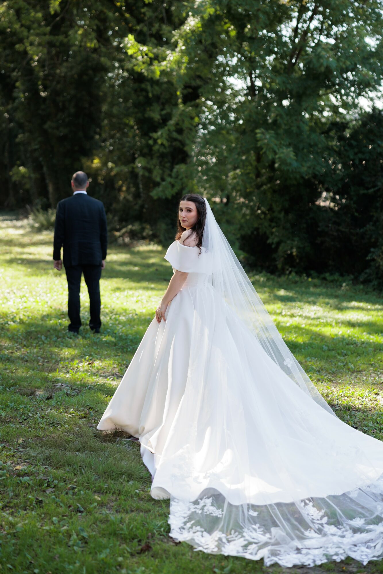 Bride in a white wedding dress and veil stands on grass, with a man in a suit facing away in the background.