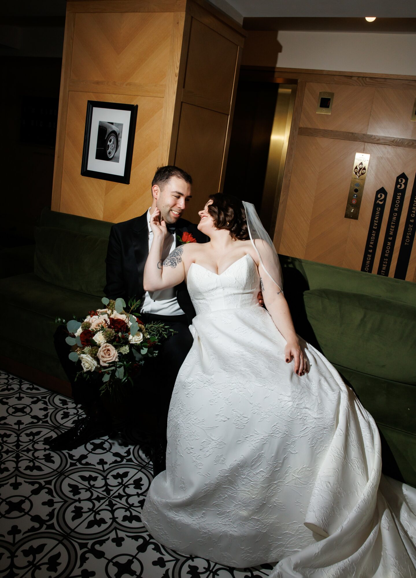 Bride and groom sitting on a green couch, smiling and touching faces, bride holding a bouquet of flowers, indoor setting.