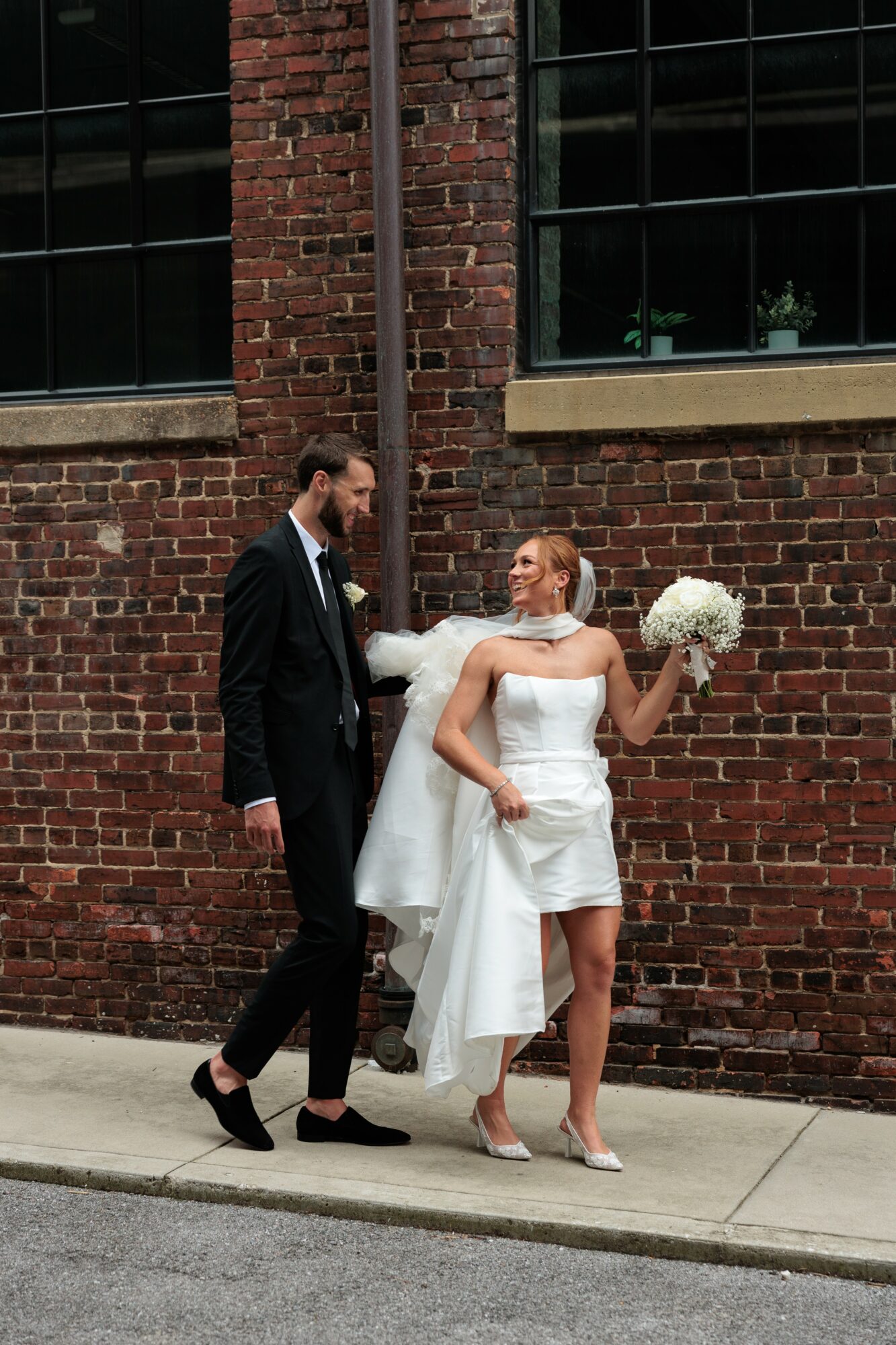 Bride in white dress holding bouquet, groom in black suit standing on sidewalk against brick wall.