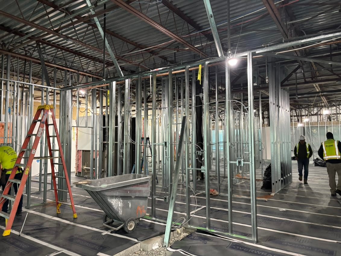 Construction site with metal framing, ladder, wheelbarrow, and two workers in safety vests.