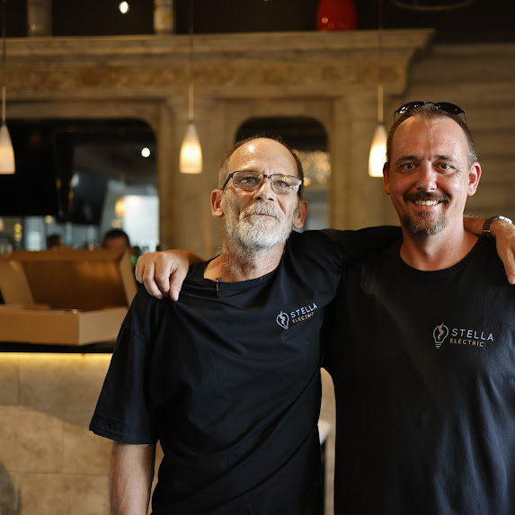 Two men in black shirts with Stella logo, smiling, standing close together indoors with warm lighting.