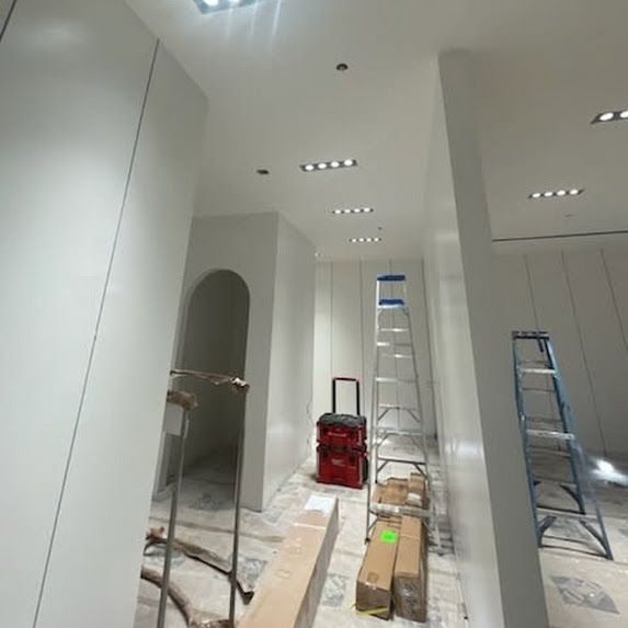 Interior space with ladders, boxes, and construction tools, white walls, ceiling lights, and an arched doorway.