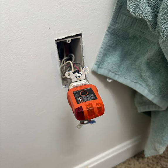 Electrical outlet with a device hanging out, wires visible, next to a towel hanging on the wall.