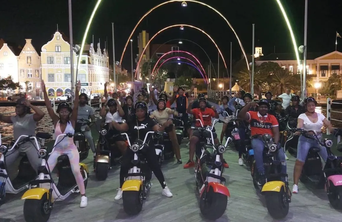 Group of people riding scooters on a street at night with colorful light arches overhead.