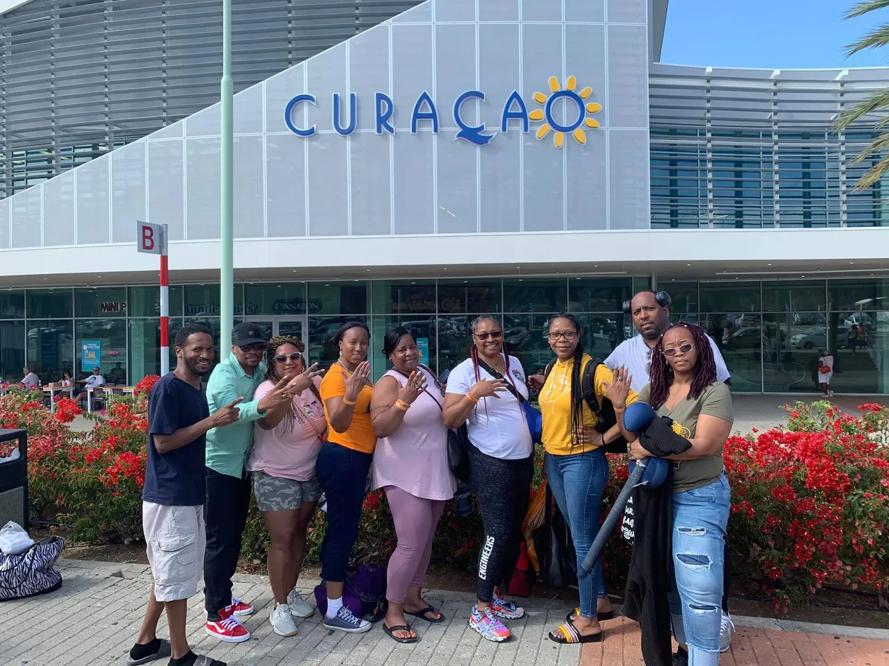 Group of eight people standing outside building with 'CURAÇAO' sign, flowers in foreground, clear sky.