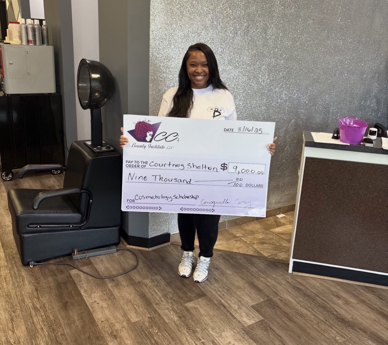 Smiling woman holding large check in a room with wooden floor and gray wall.