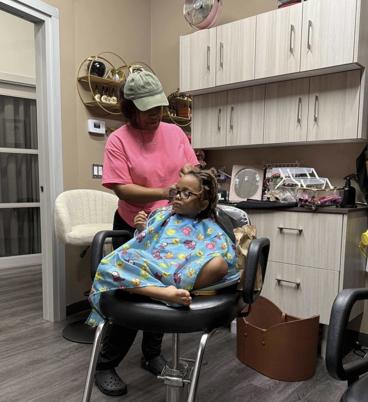 Person with glasses and colorful cape sitting in a salon chair, another person standing behind, in a room with cabinets and kitchen items.