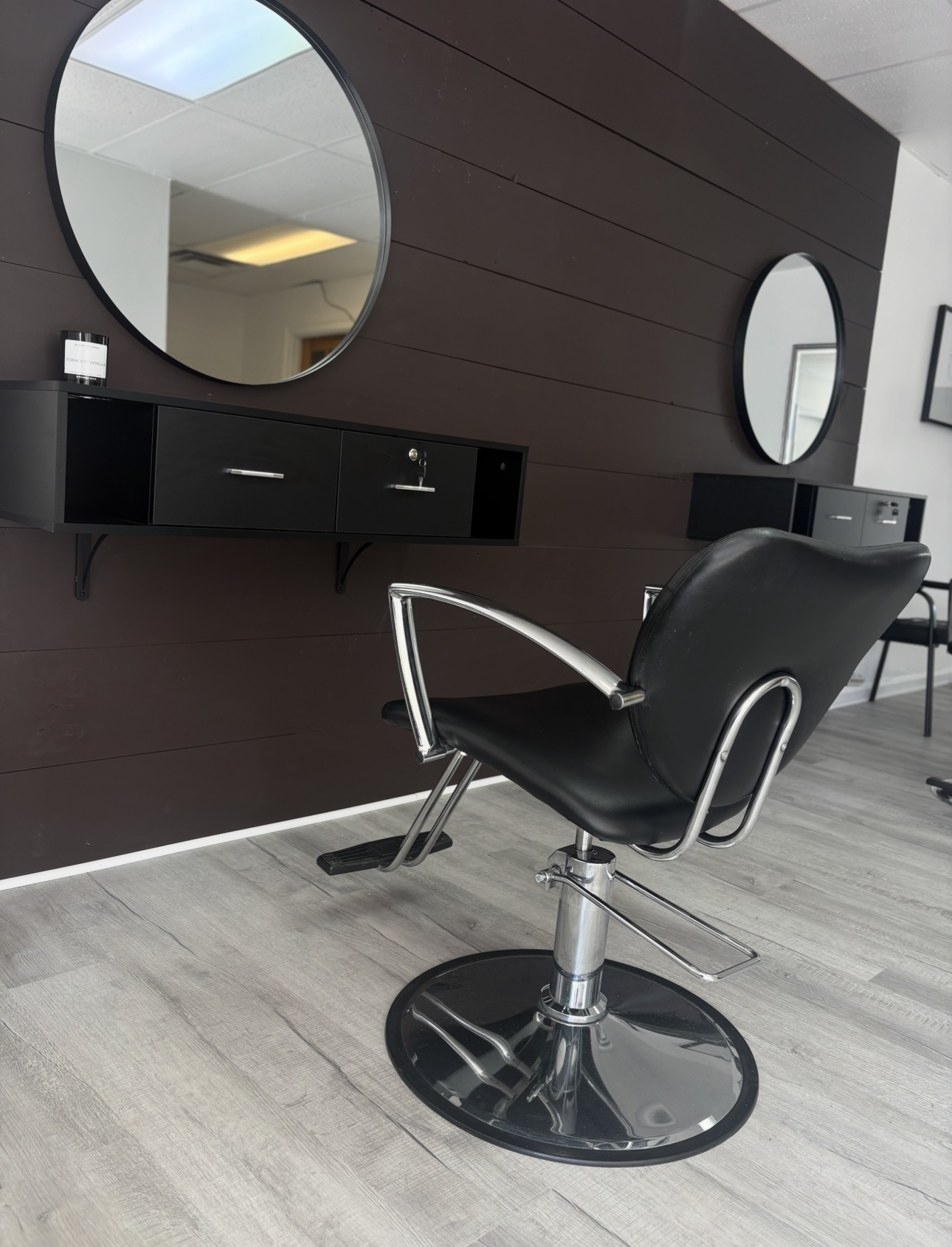 Salon chair in front of dark wood wall with two round mirrors and a black shelf.