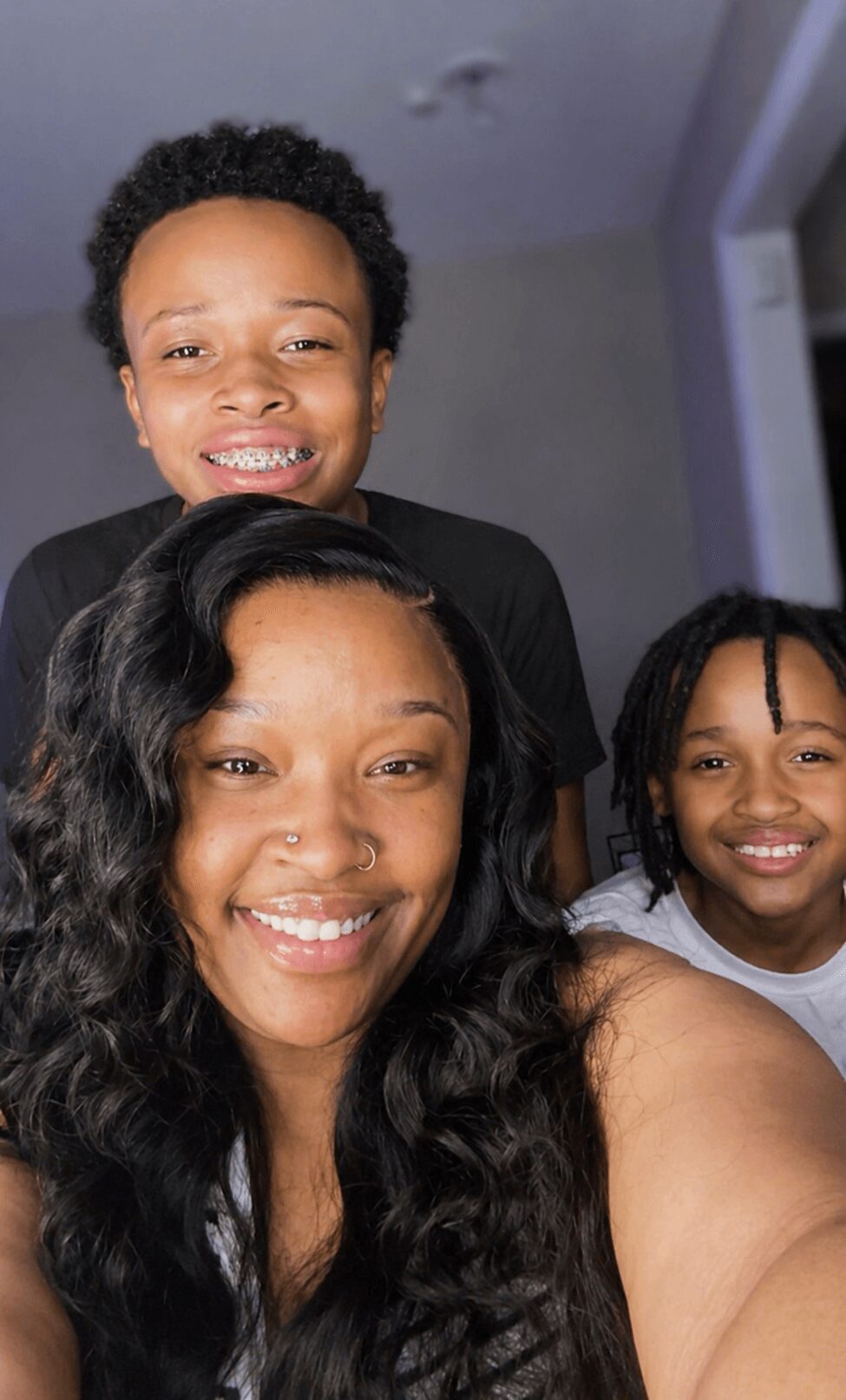 Three smiling people, two children and an adult woman, indoors with a neutral background.