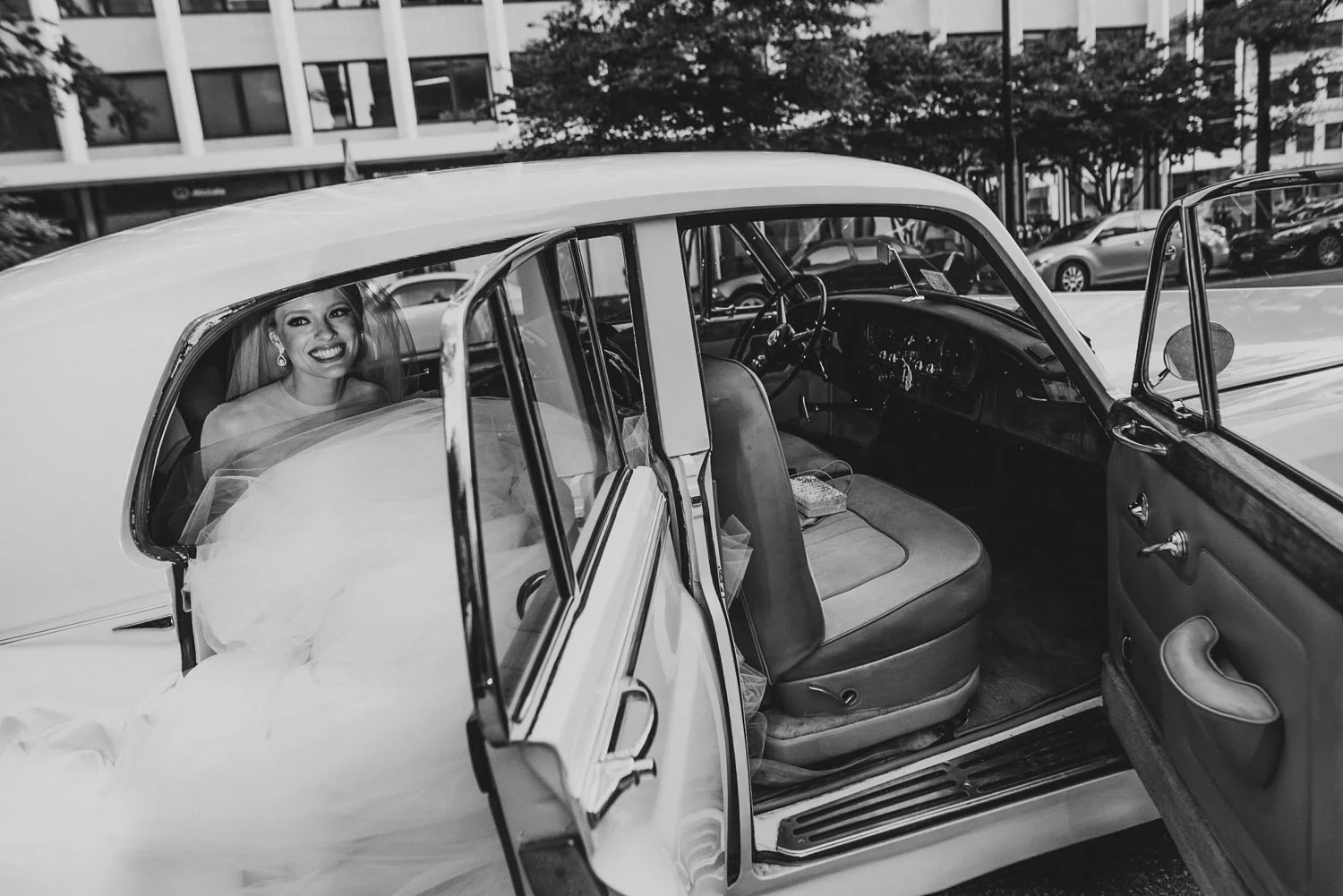 Black-and-white photo of a smiling bride sitting inside a vintage car outside The St. Regis in Washington DC, capturing an elegant and timeless luxury wedding moment.