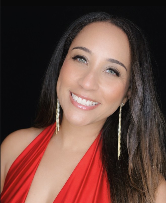 Smiling woman with long dark hair, wearing a red dress and long earrings, against a black background.