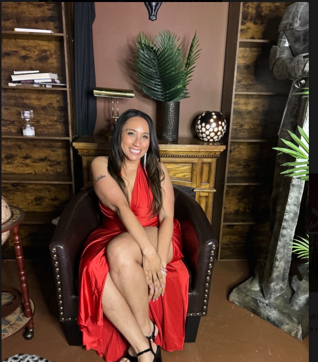 Woman in red dress sitting on a dark armchair, smiling, with wooden shelves, plants, and a statue in the background.