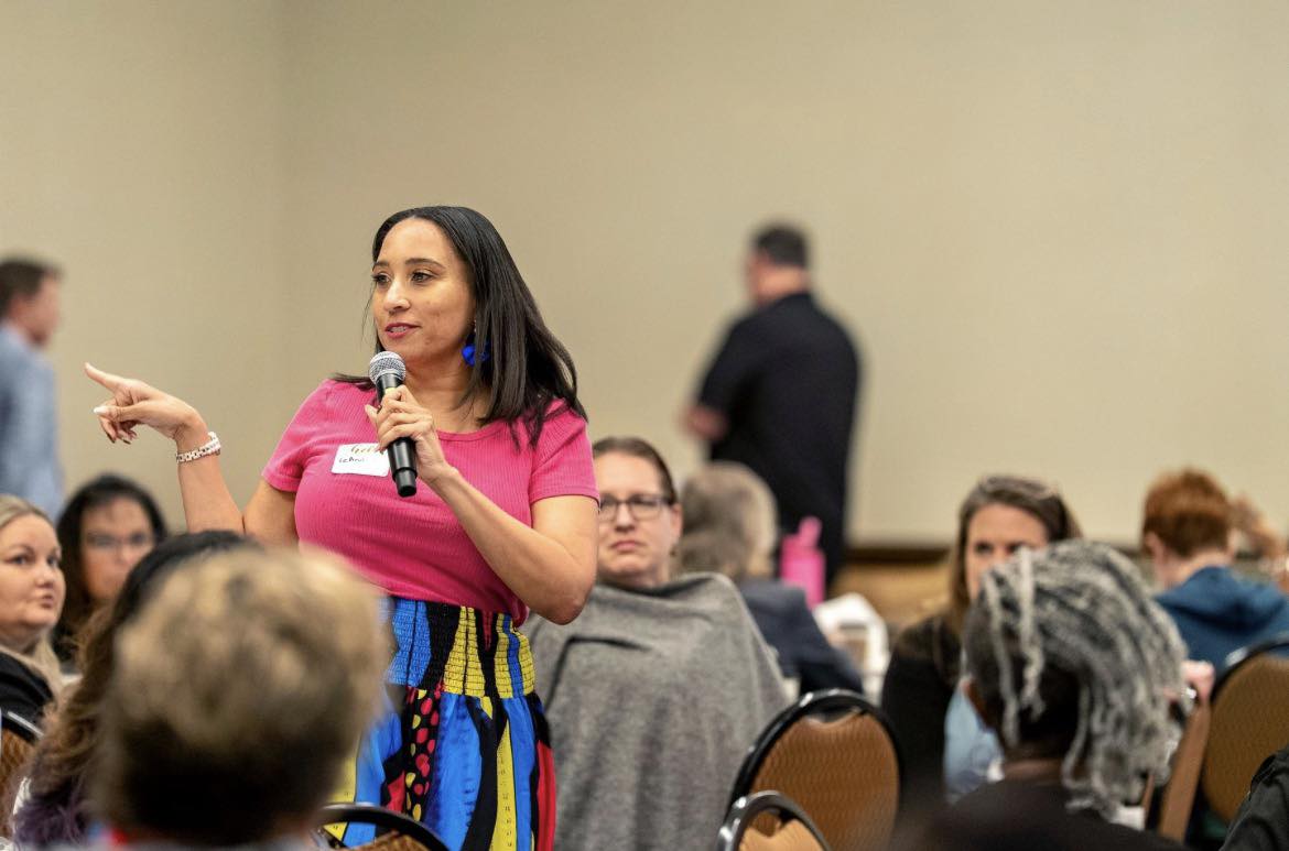 Woman in pink shirt speaking into microphone at a conference, gesturing with her left hand, audience seated around her.
