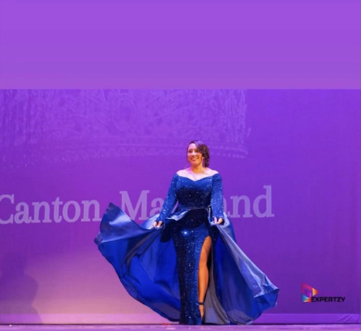 Woman in a blue gown with a high slit, standing on stage with purple background and text, holding the gown's sides.