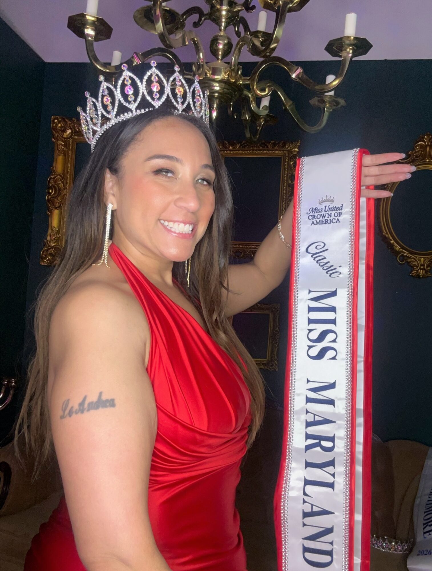 Woman wearing a crown and red dress holding a sash that reads 'Miss Maryland.' She is smiling and standing indoors.