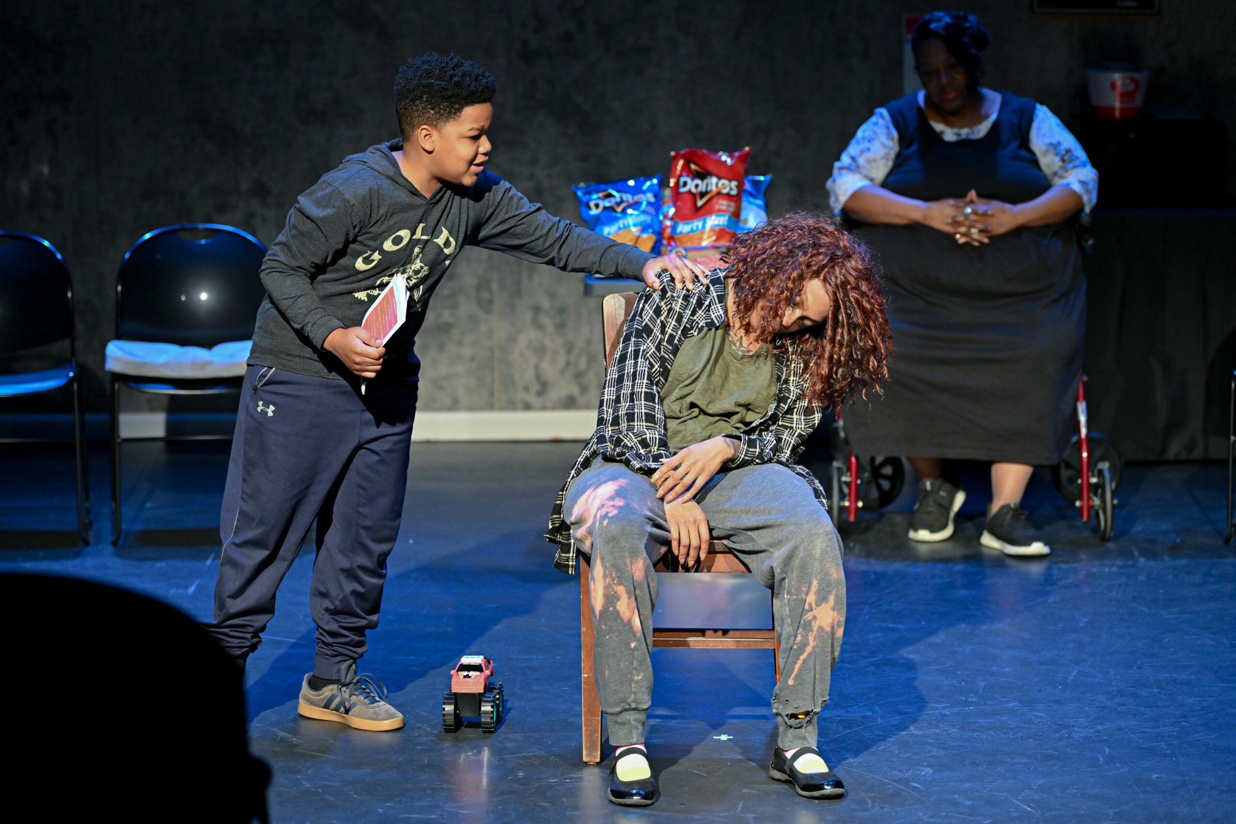 A young boy interacts with a seated person with curly hair on stage, with a woman in the background. Stage setting with chairs.