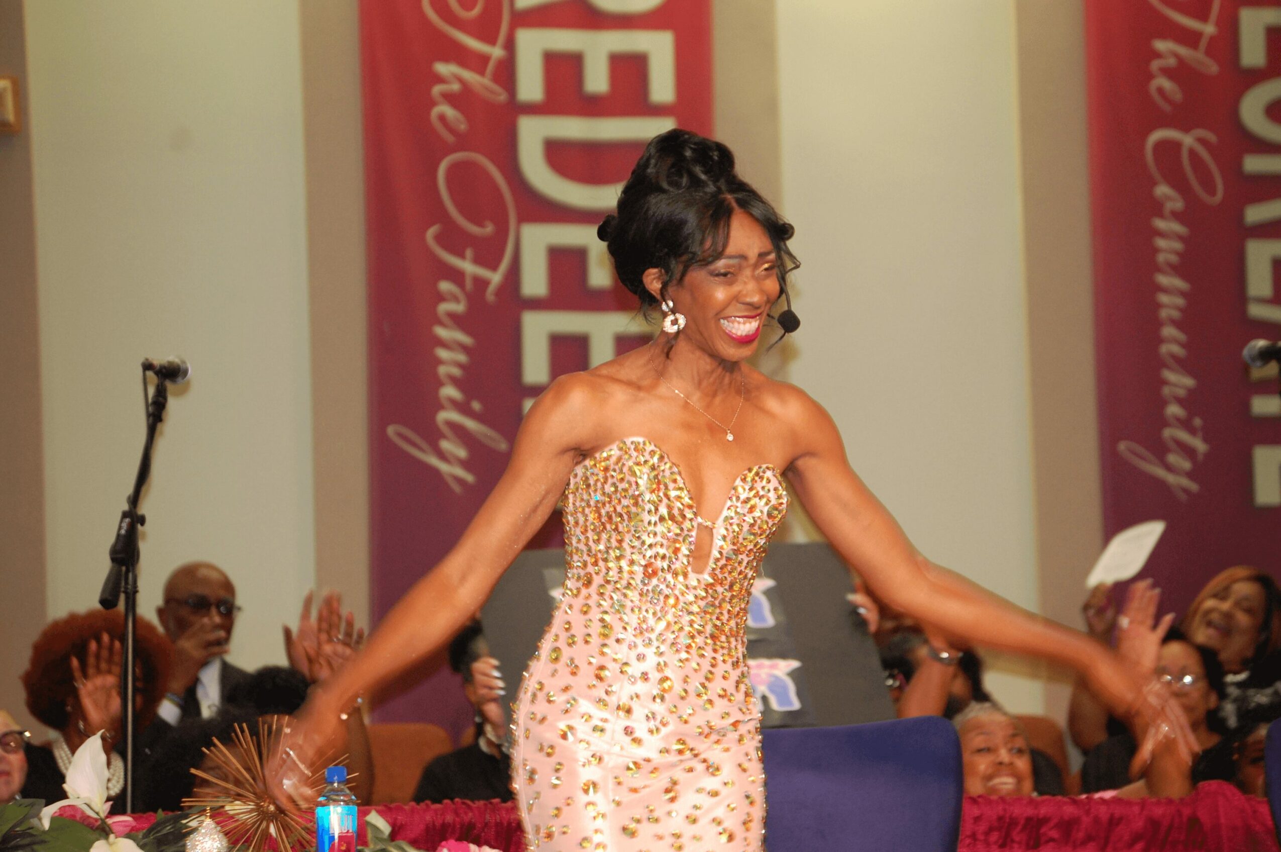 Woman in a sparkling, strapless gown smiling and dancing at an event with seated audience and banners in background.