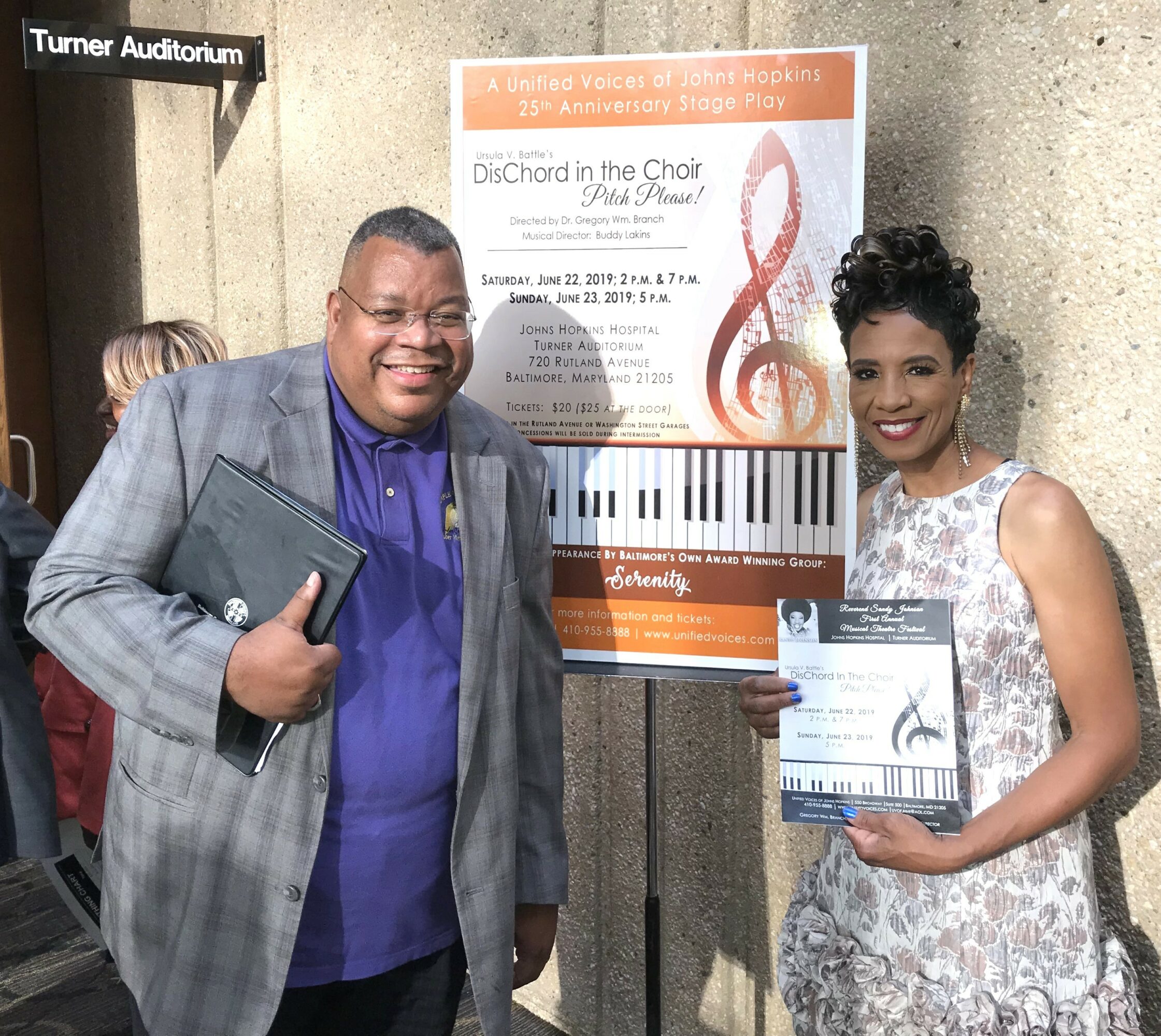 Two people standing in front of a poster, smiling, holding certificates, in a building lobby with a sign reading 'Turner Auditorium'.