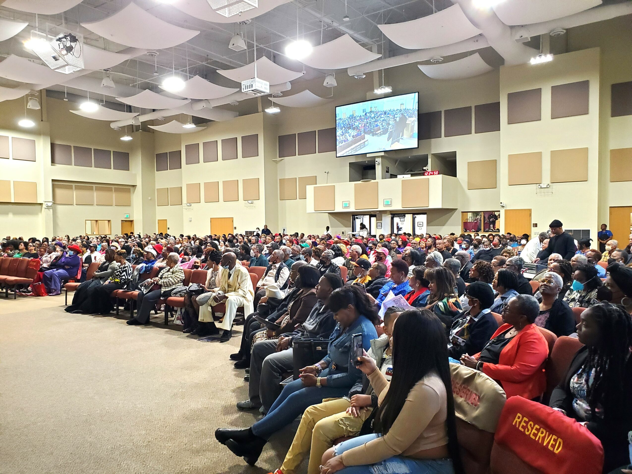 Large indoor gathering with many seated people facing a stage with a large screen, in a spacious room with high ceiling.