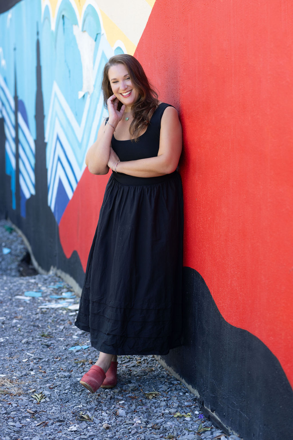 Woman in black dress and pink shoes standing against a colorful graffiti wall, smiling, with arms crossed.