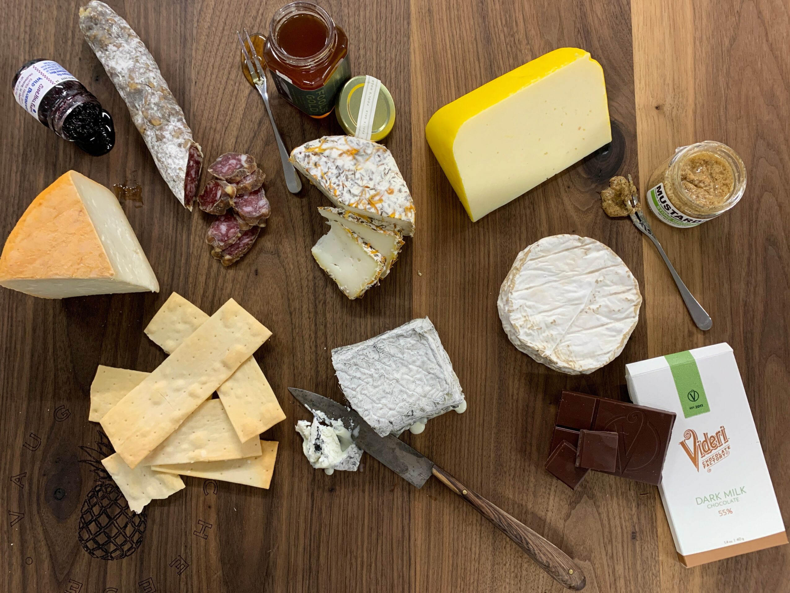 Assorted cheeses, honey, jam, and crackers on a wooden surface, with a knife and a small glass of beverage.