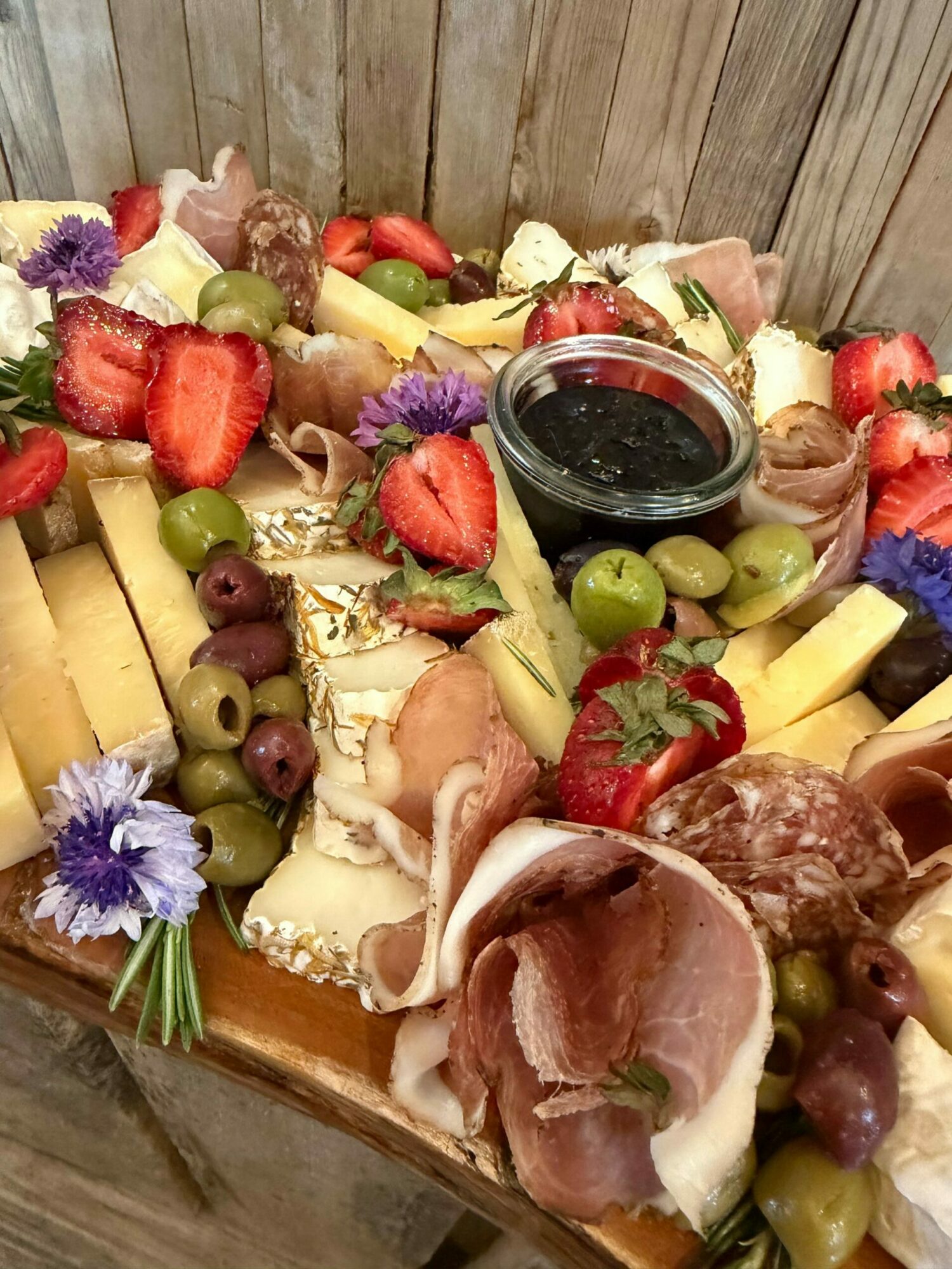 Assorted cheese, grapes, strawberries, cured meats, and flowers on a wooden board.