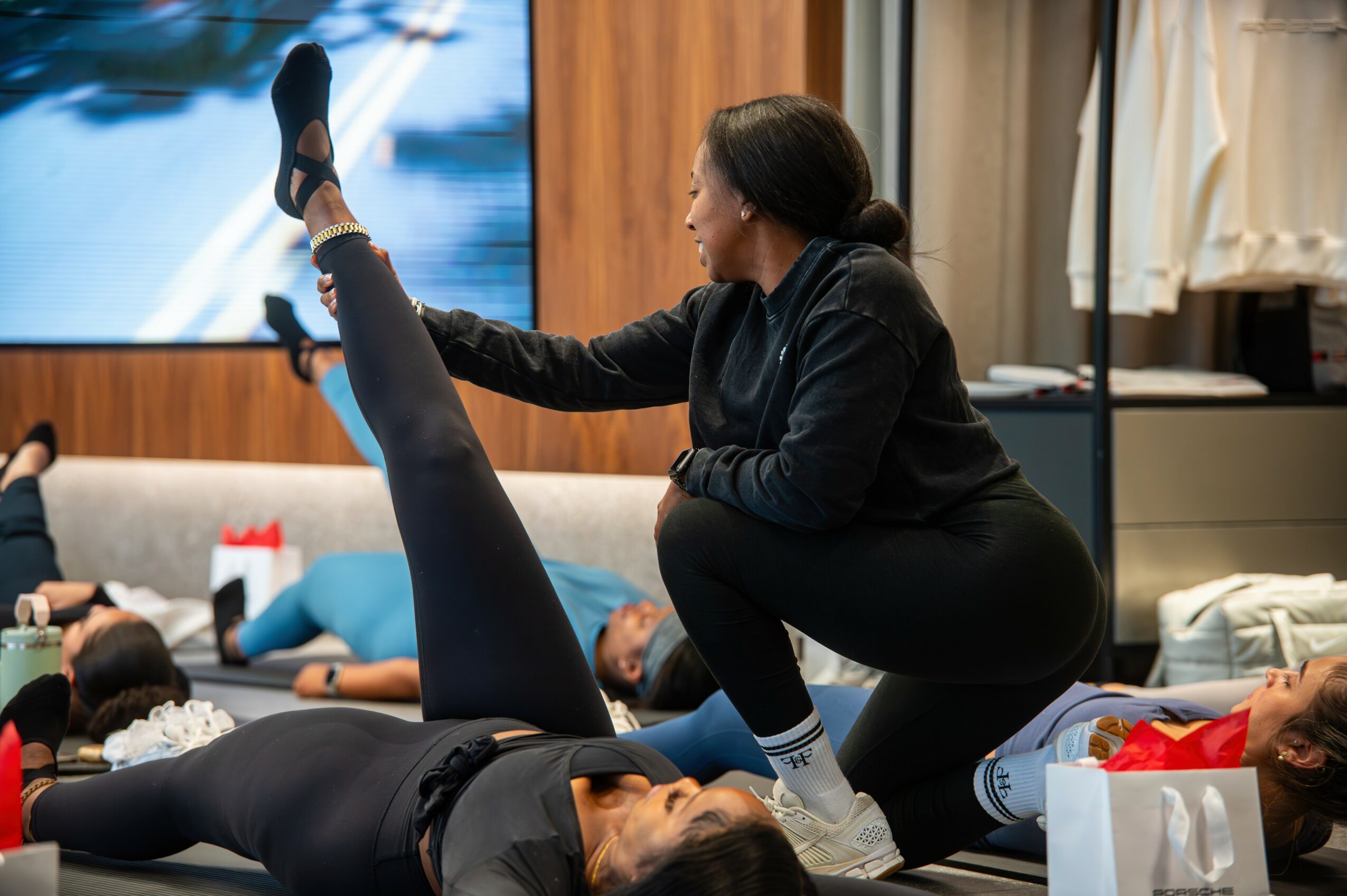 Instructor assisting a person with stretching exercises in a fitness class, with others lying on mats in the background.