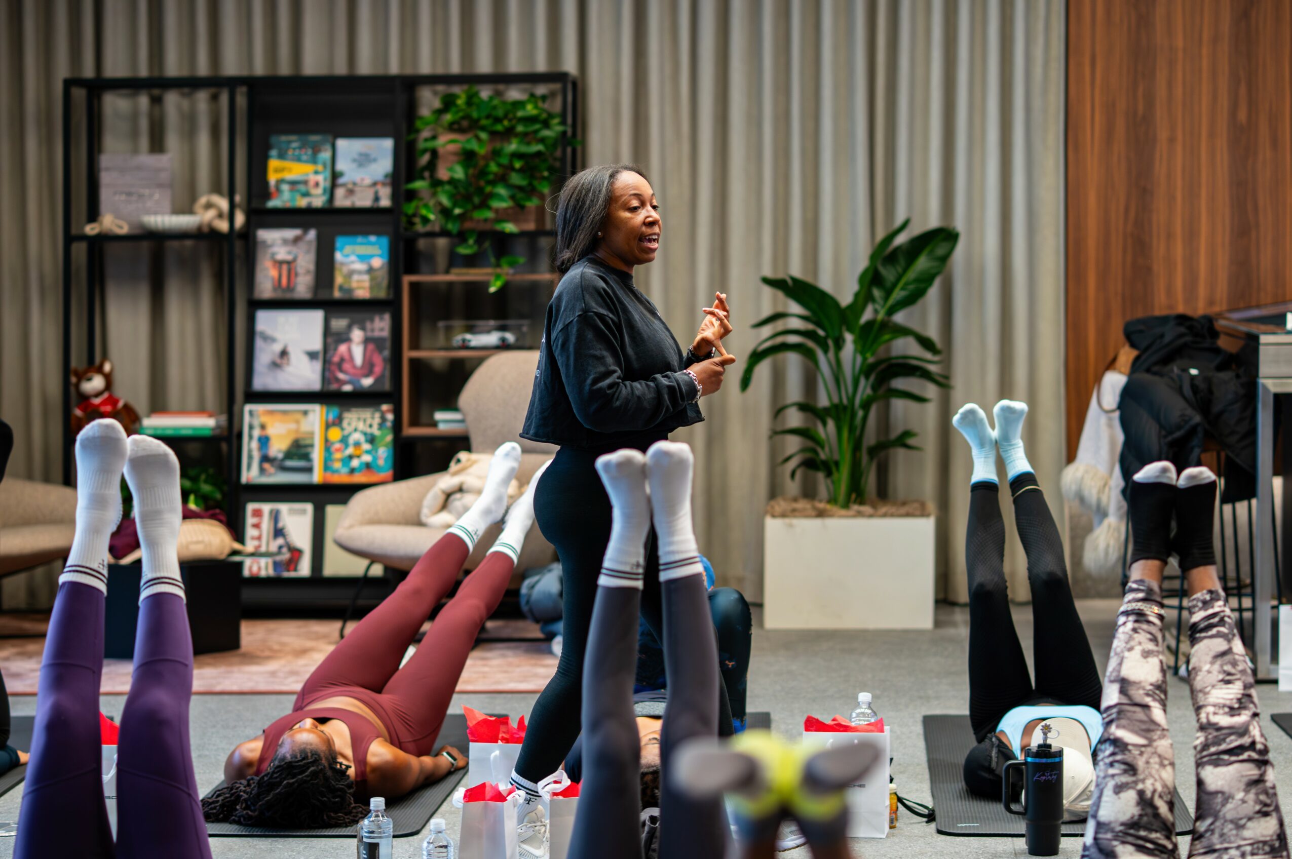 Woman leading a fitness class with people lying on their backs with legs raised, in a room with bookshelves and plants.