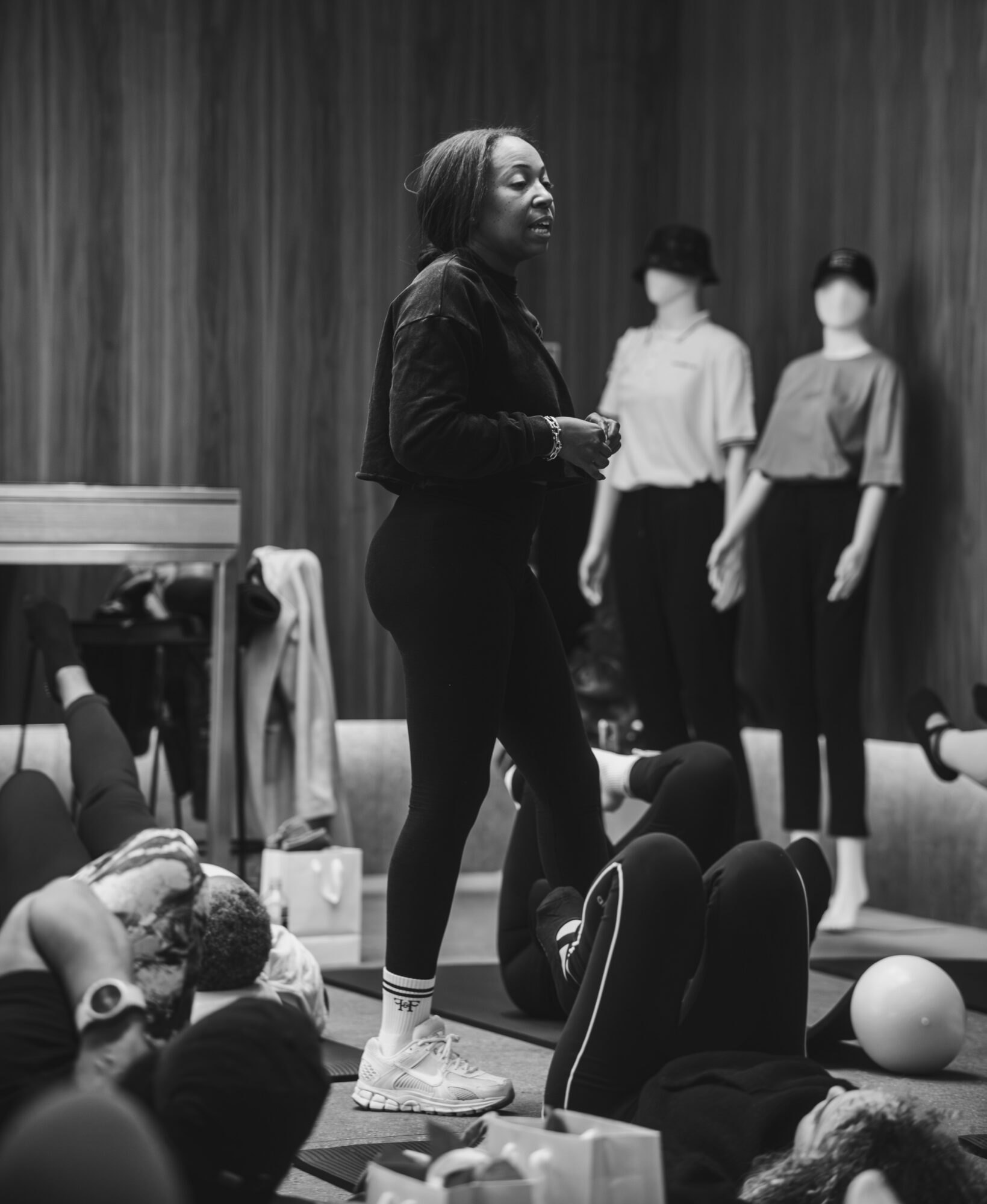 Woman standing and speaking to a group of people sitting on the floor, with two women standing in the background, all in a room with a curtain backdrop.