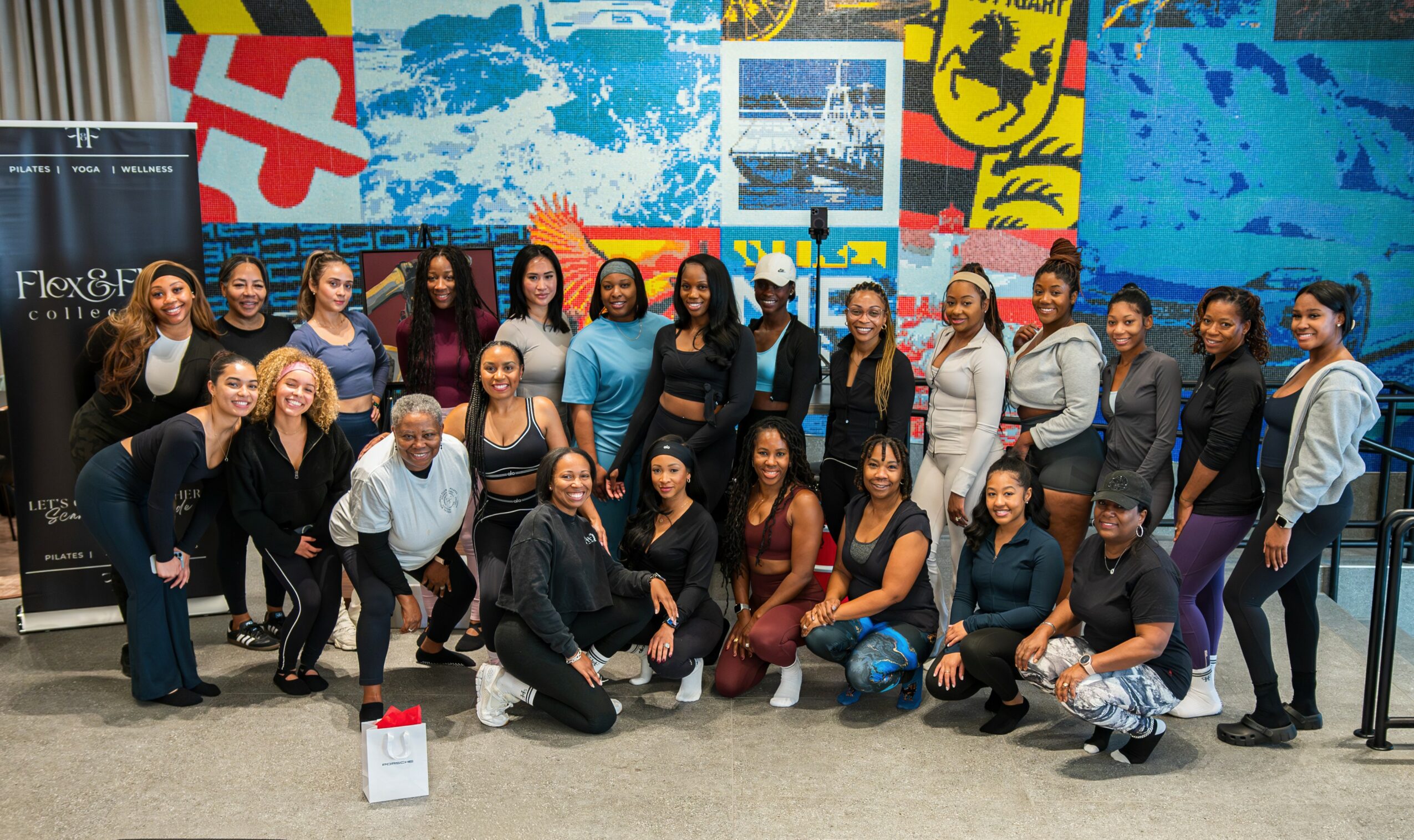 Group of diverse people posing indoors in front of colorful wall art, some kneeling and others standing, smiling.