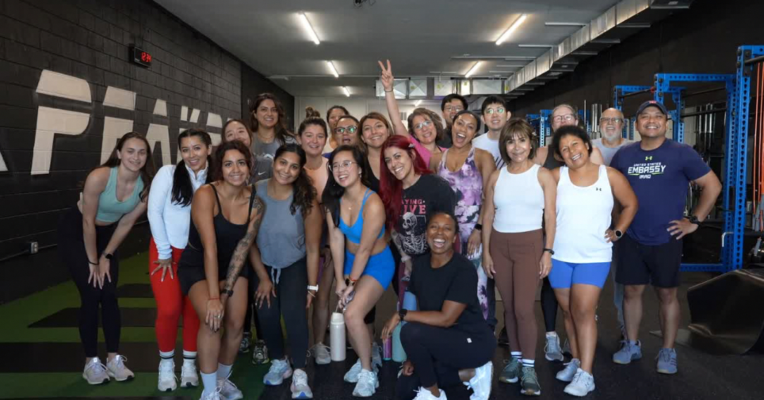 Group of diverse people in workout attire posing indoors, some smiling and making gestures, gym setting in background.