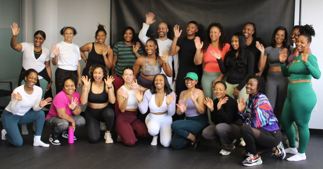 Group of diverse women smiling and waving in front of a black backdrop, some kneeling and some standing.