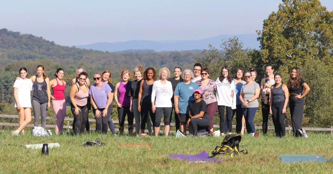Group of people outdoors on grass, some with yoga mats, with trees and hills in background.