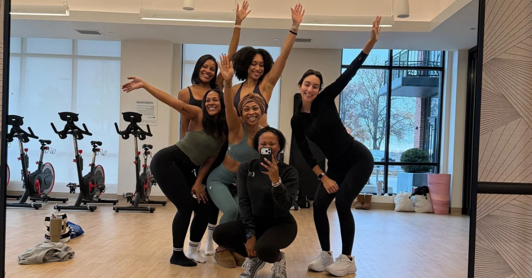 Six women in workout clothes pose in a fitness studio, some with arms raised, smiling, with exercise bikes in background.