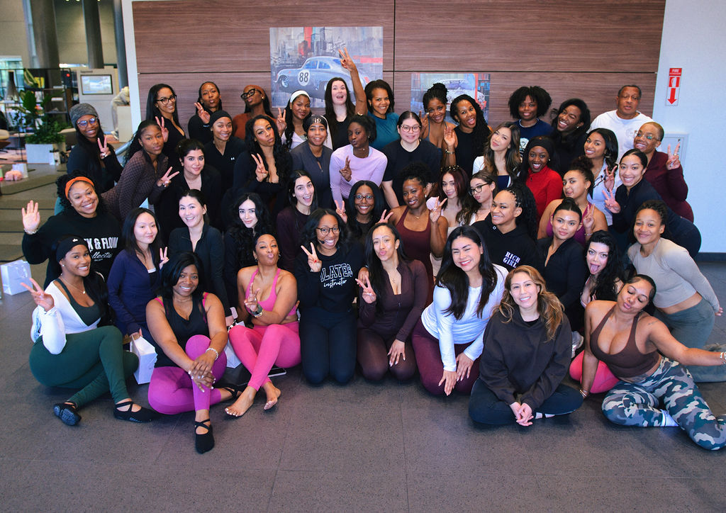 Group of diverse women and men posing together indoors, some sitting and some standing, smiling at the camera.
