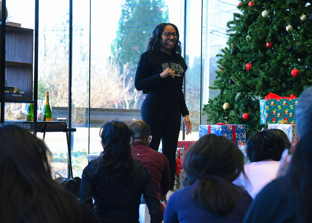 Woman speaking to an audience during a holiday event with a decorated Christmas tree and presents, large windows in background.