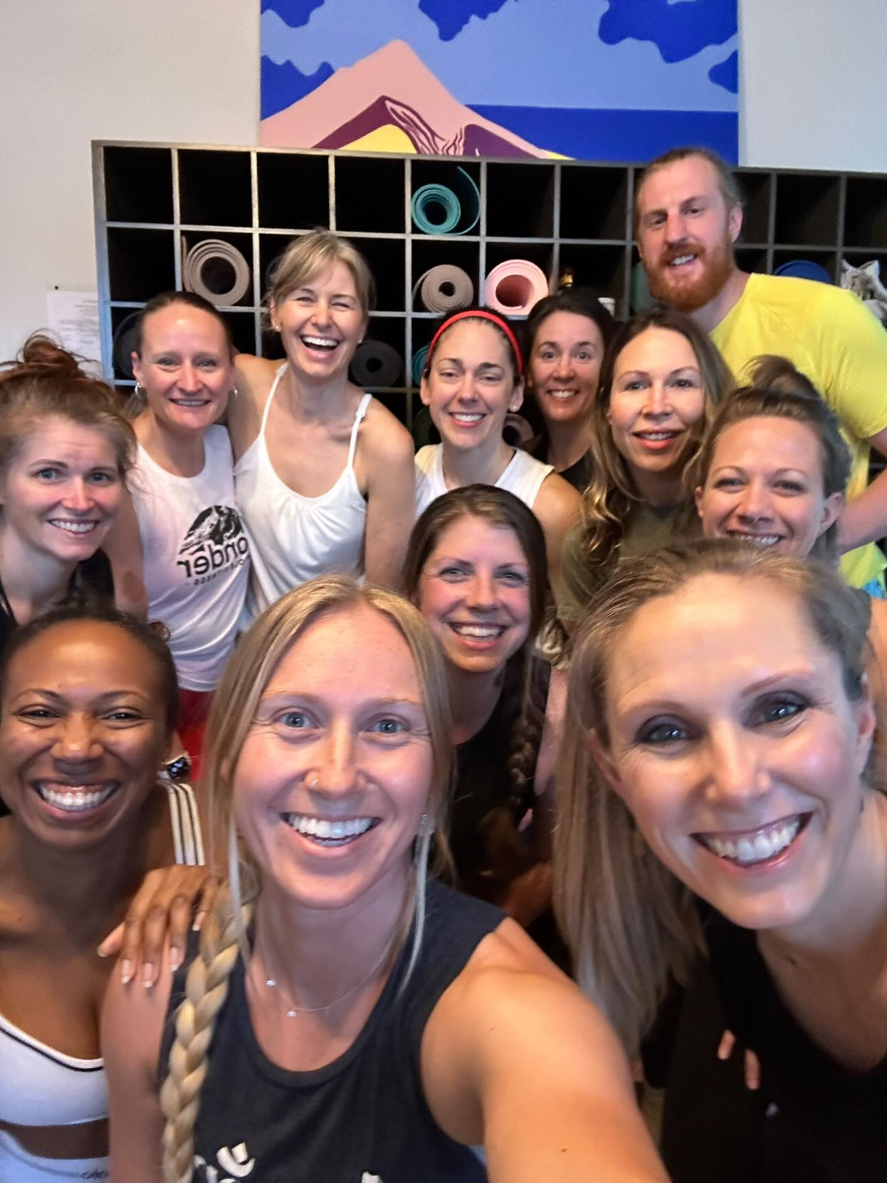 Group of fifteen smiling people taking a selfie indoors, with a colorful mural and shelves in the background.