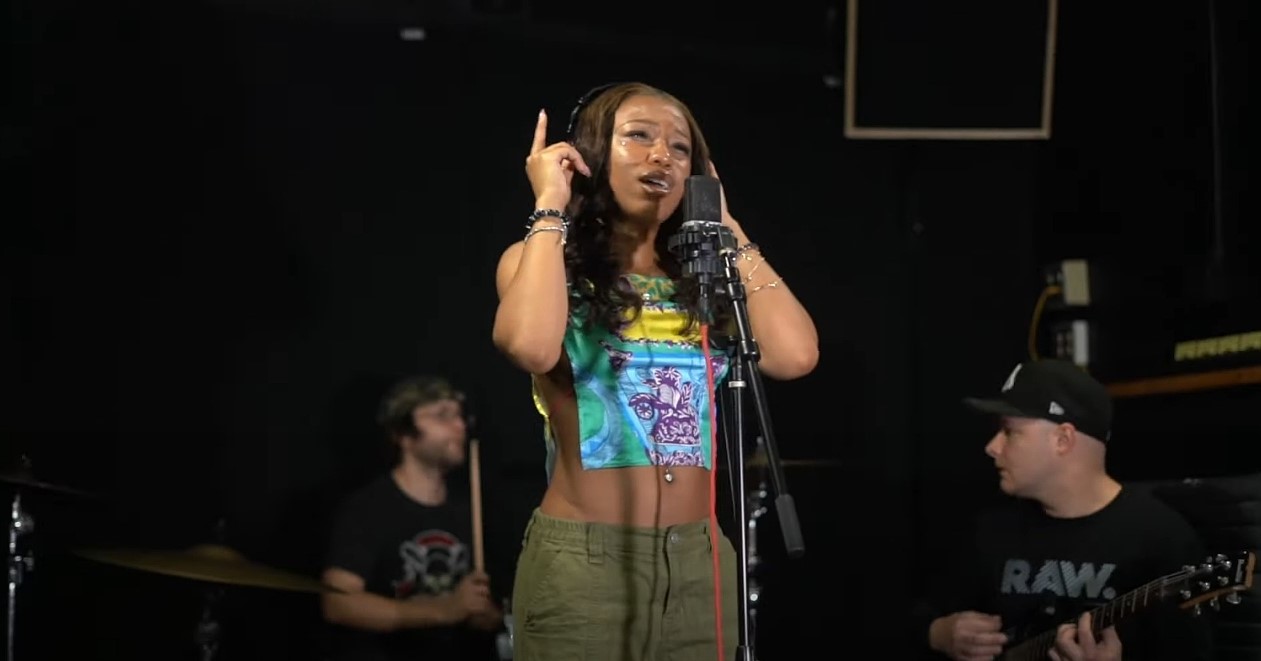 A woman sings into a microphone with two men playing instruments in the background, in a dark studio or stage setting.