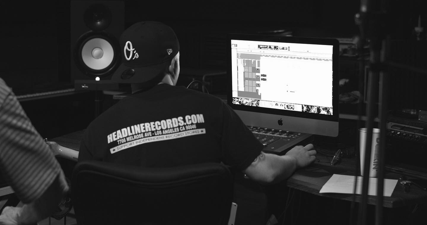 Person working at computer in a music studio with audio equipment, wearing a cap and a T-shirt with a website URL.