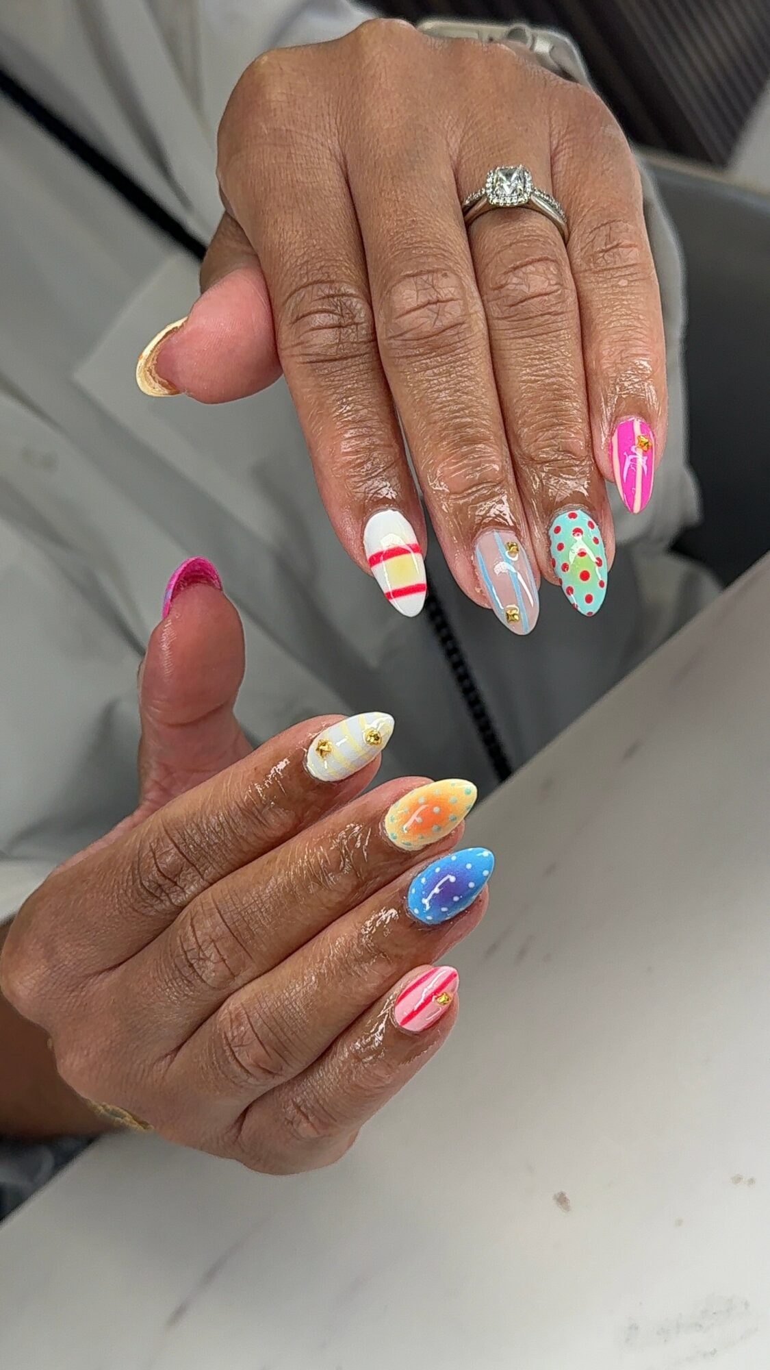 Two hands with colorful, decorated nails resting on a surface, one hand with a ring, nails with various patterns and colors.
