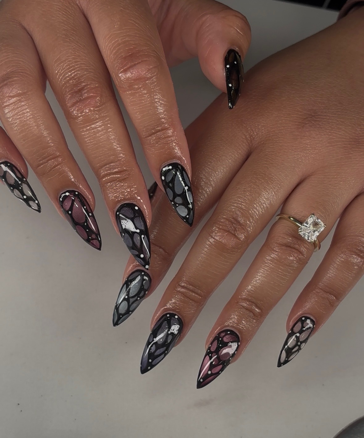 Two hands with long, pointed nails painted in black, pink, and gray with geometric patterns, one hand has a ring.