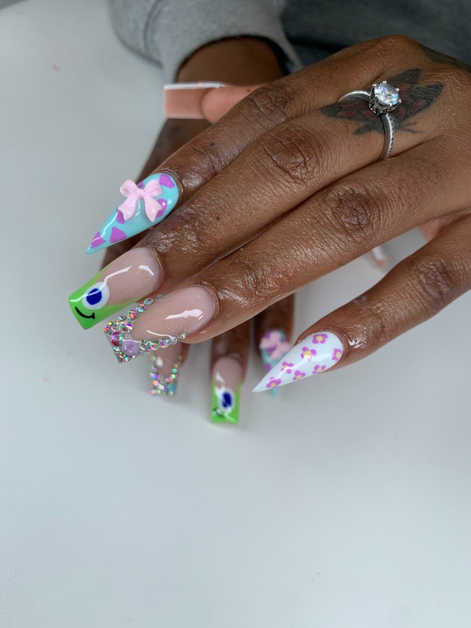 Close-up of hands with decorated nails, including a ring with a large gemstone, and a tattoo on the finger.