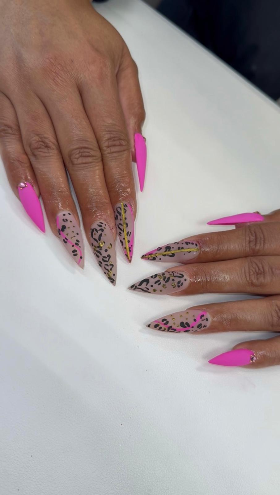 Two hands with long, pointed nails painted bright pink, featuring leopard print and pink accents, resting on a white surface.