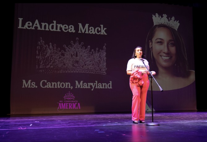 Person standing on stage with a microphone, large screen behind displaying a smiling woman's face and text, stage lighting.